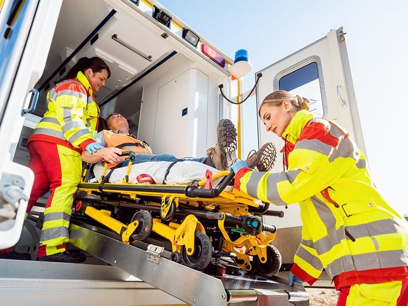 Paramedics load an injured patient into an ambulance using a wheeled stretcher and ramp system.