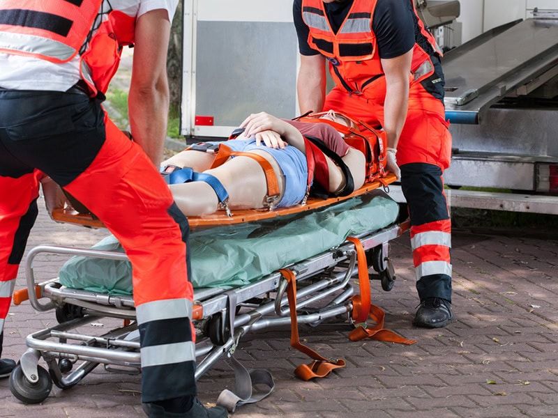 Emergency personnel transfer an injured patient secured on a spine board onto a wheeled stretcher for ambulance transport.